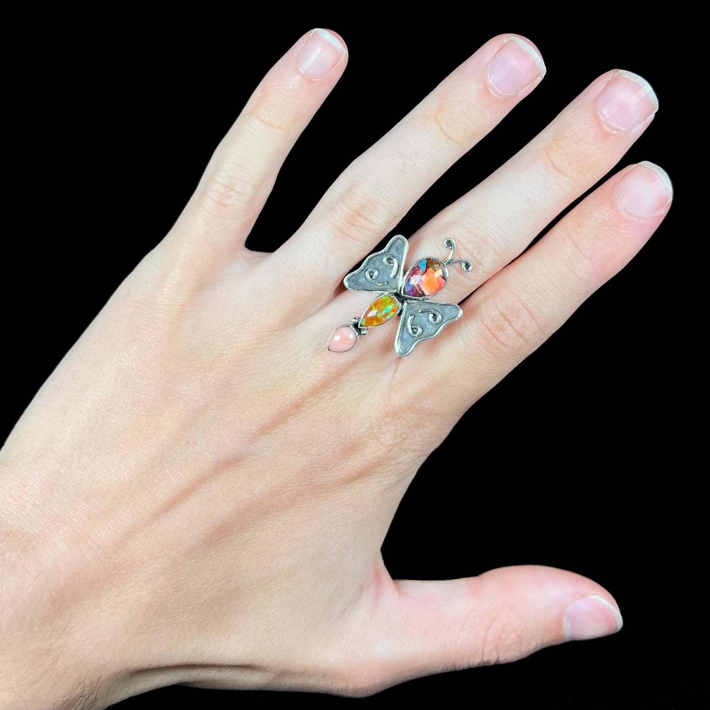 A silver multi-gemstone butterfly ring worn on a hand against a black background.