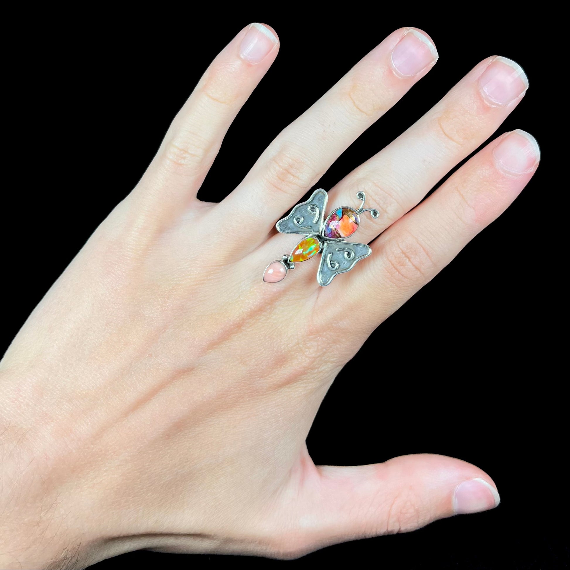 A silver multi-gemstone butterfly ring worn on a hand against a black background.