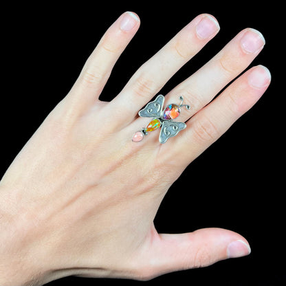 A silver multi-gemstone butterfly ring worn on a hand against a black background.