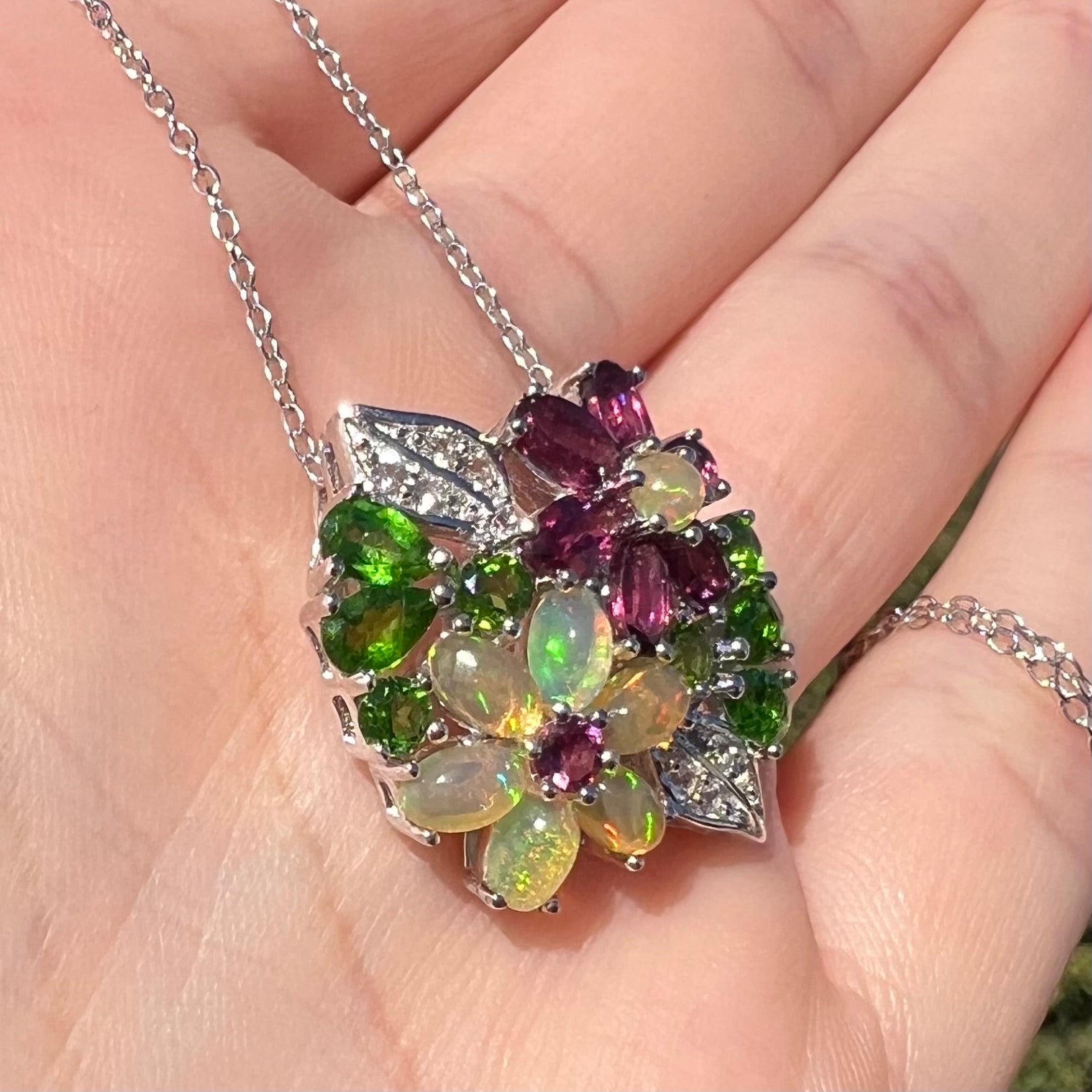 A sterling silver opal and garnet flower necklace shown close-up in the palm of a hand at an angle in sunilght.