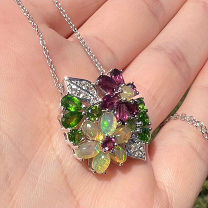 A sterling silver opal and garnet flower necklace shown close-up in the palm of a hand at an angle in sunilght.