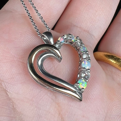 Close-up angled view of a sterling silver and yellow gold lab-created opal and CZ heart necklace in a hand.