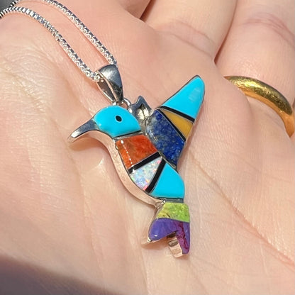 A multi-stone inlay hummingbird necklace in sterling silver, shown close-up at an angle in a hand in sunlight.