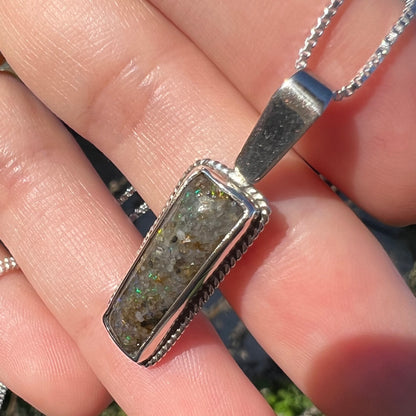 An angled close-up of a Louisiana matrix opal pendant in sterling silver shown in the sunlight, held in a hand.