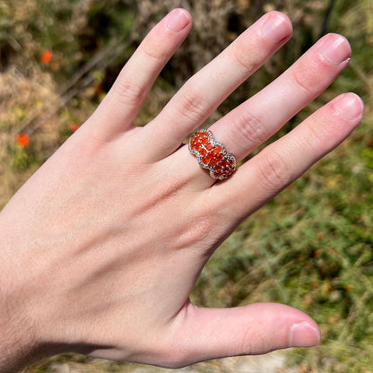 Hand in sunlight wearing a sterling silver Mexican fire opal and diamond-accent ring against blurred green outdoor background.  The many faceted round opals are orange.