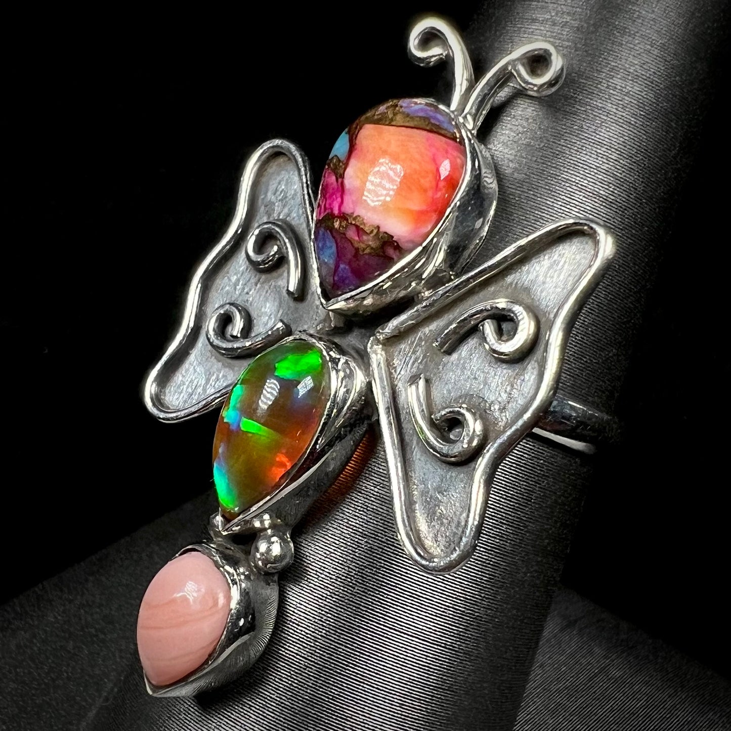A silver opal butterfly ring shown at an angle against a black background.