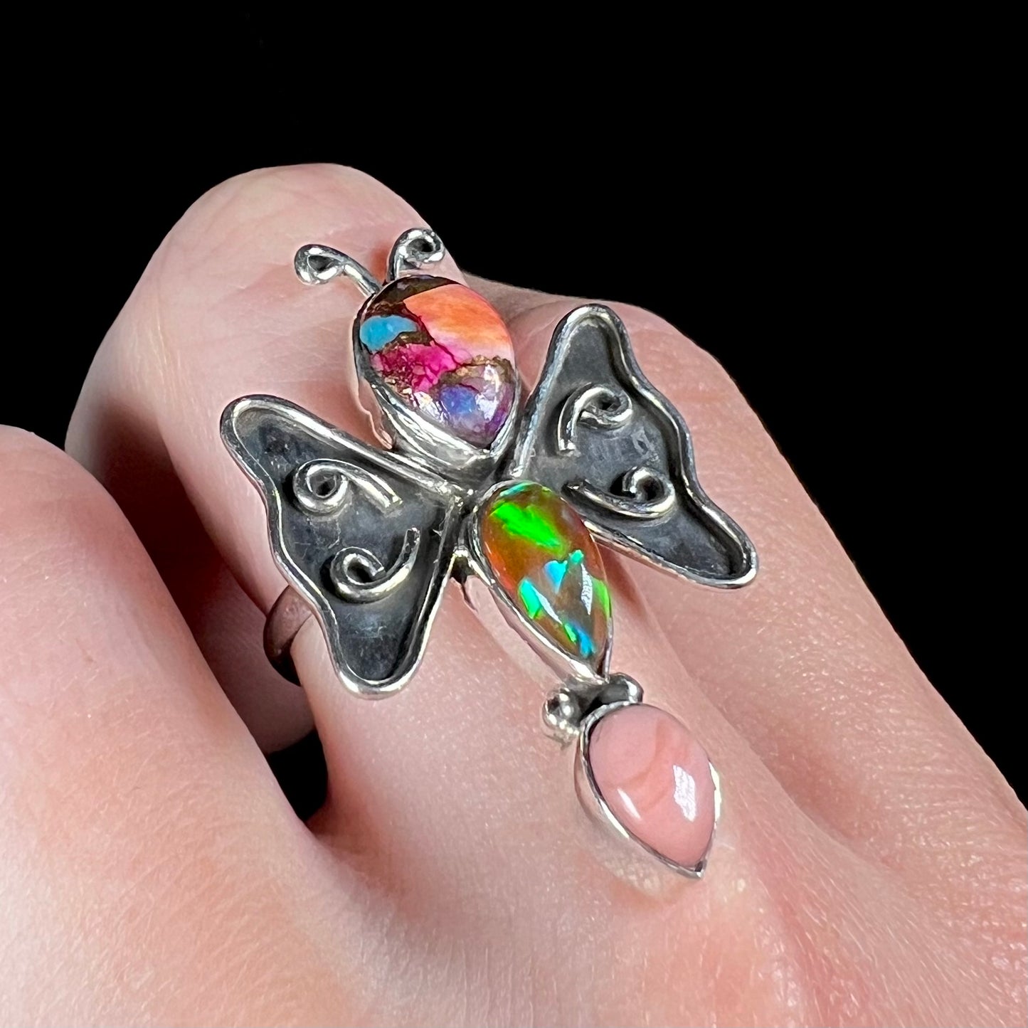 A silver opal butterfly ring shown at an angle on a finger; the lab-created fire opal plays green and blue colors.