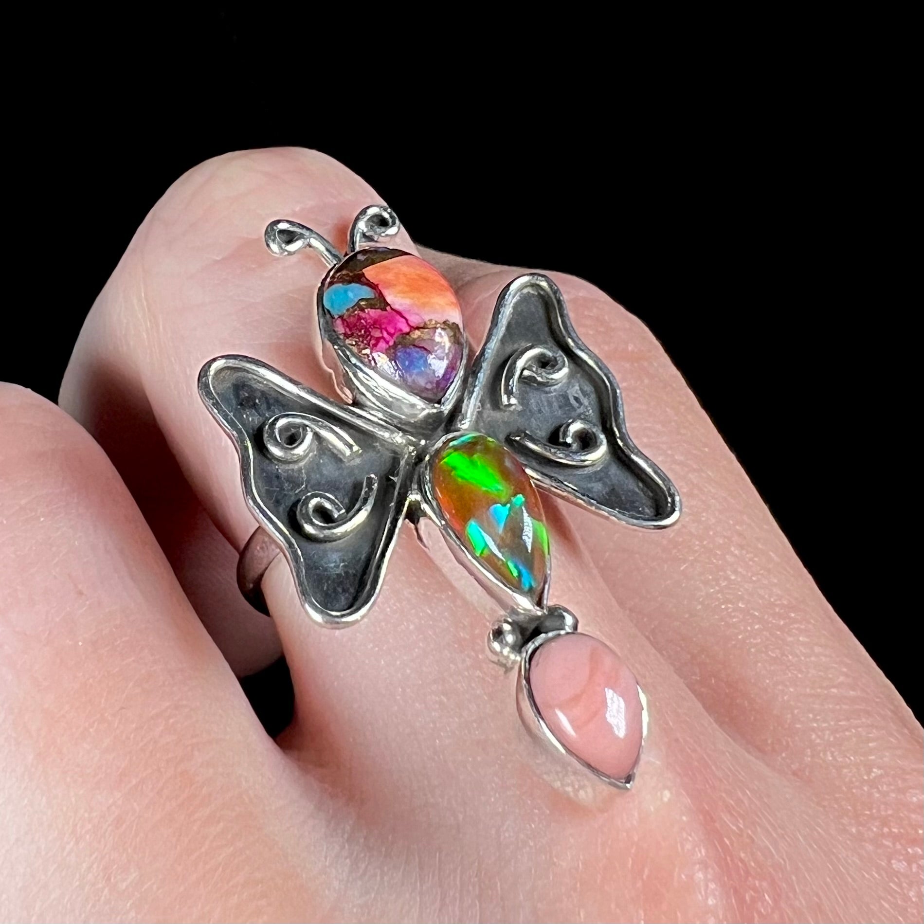 A silver opal butterfly ring shown at an angle on a finger; the lab-created fire opal plays green and blue colors.