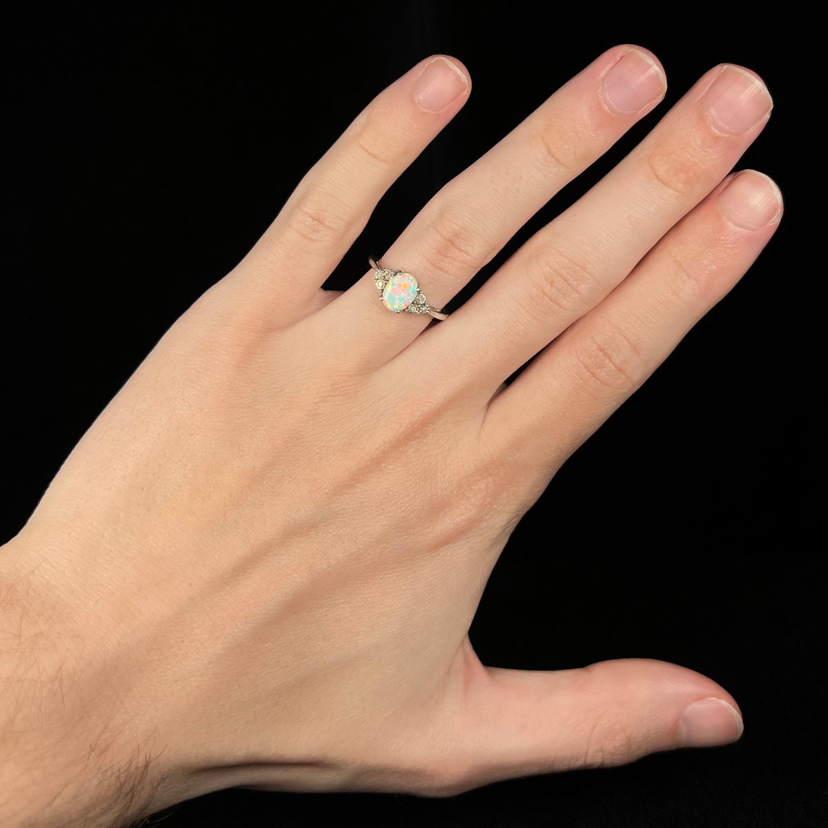 Hand wearing a sterling silver lab-created opal and diamond engagement ring.  The opal has red play-of-color.