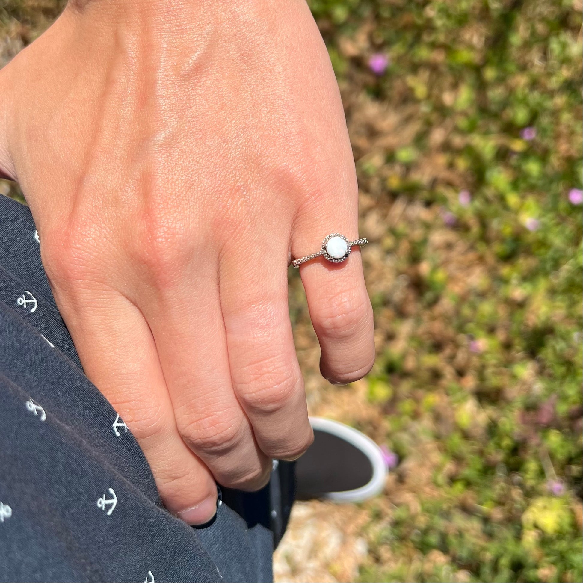 A silver opal and diamond halo ring worn on the pinky finger of a hand as it rests on the model's hip.