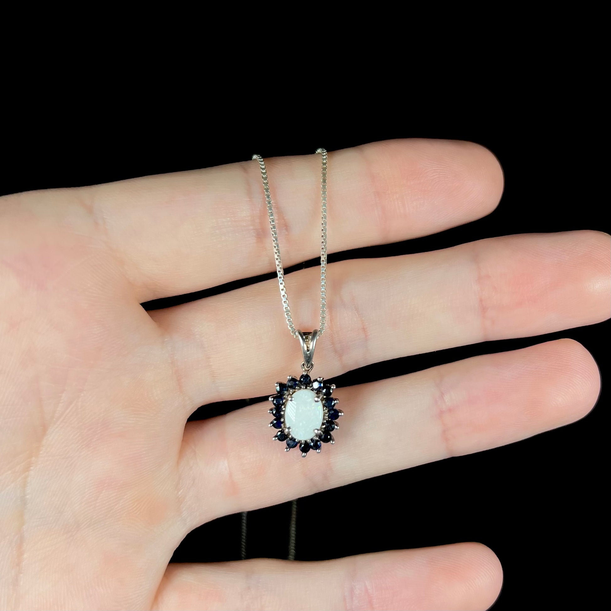 A sterling silver opal halo necklace resting on fingers against a black background.