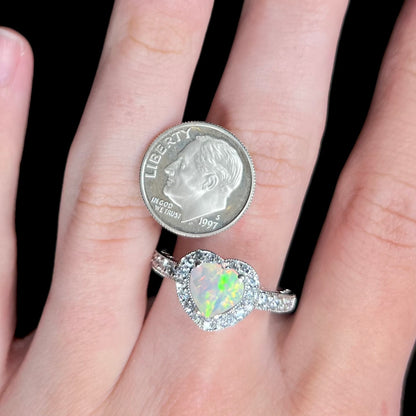 Close-up of hand wearing sterling silver opal heart ring with halo design next to US dime coin for scale