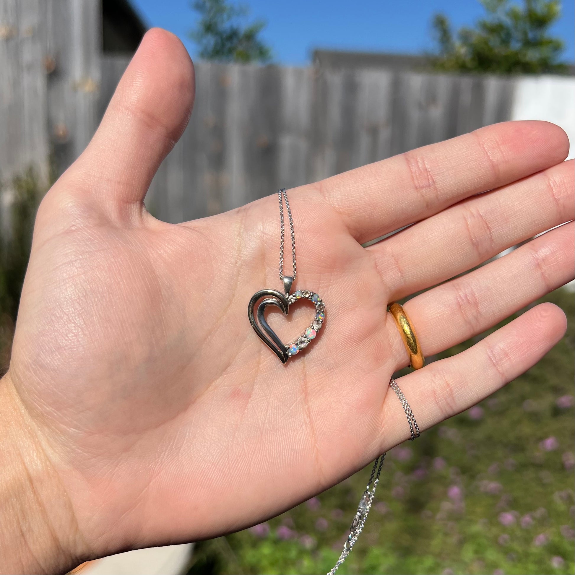 A two-tone sterling silver and yellow gold lab-created opal and CZ heart necklace in a hand in sunlight.