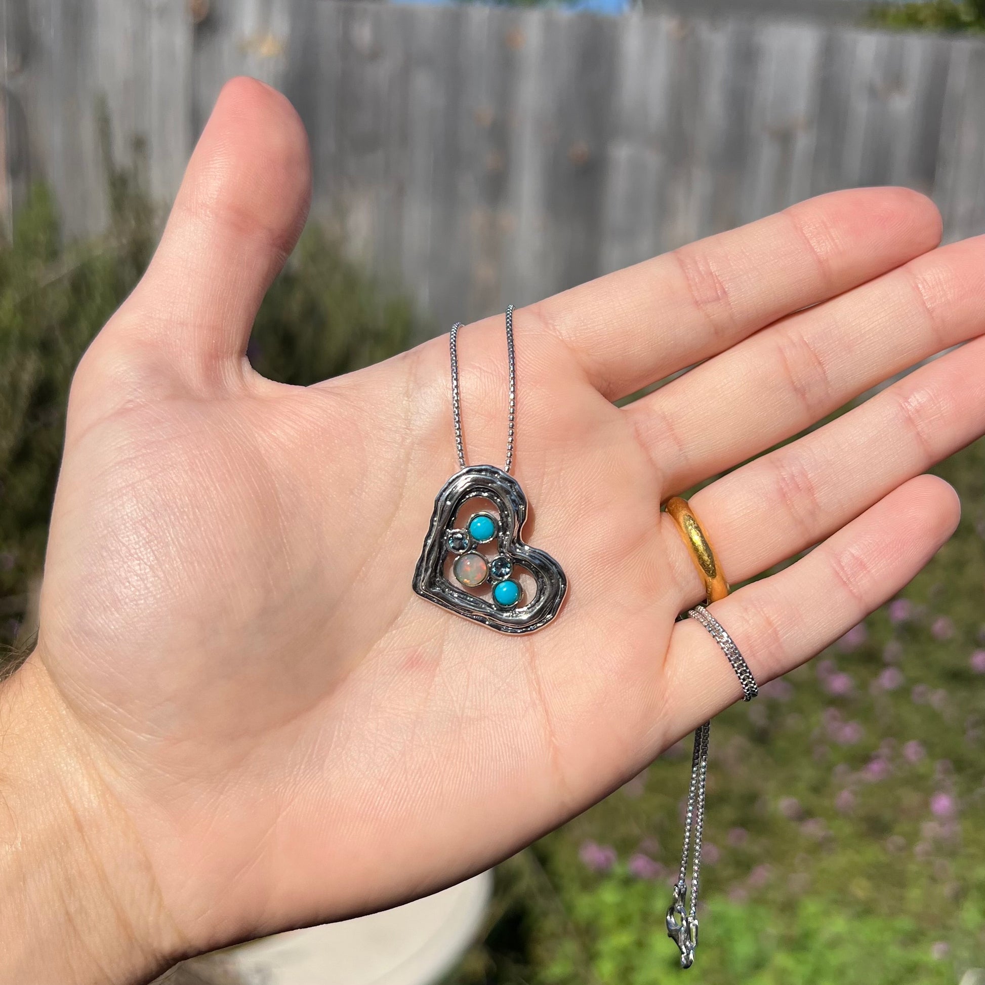 A sterling silver heart necklace set with opal, turquoise, and blue topaz, shown in a hand in outdoor sunlight.