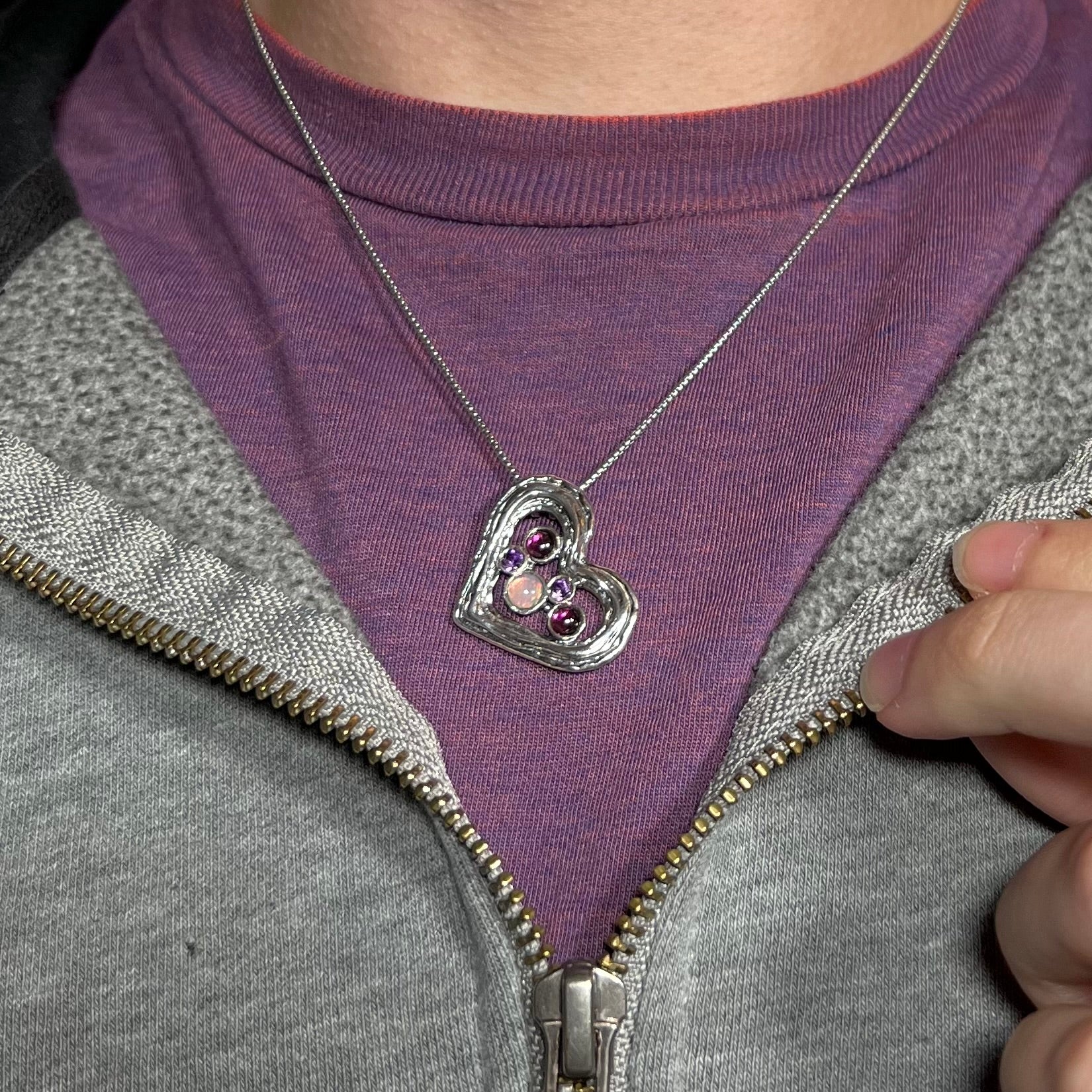 A sterling silver heart necklace with opal, amethysts, and garnets, worn against a purple shirt.