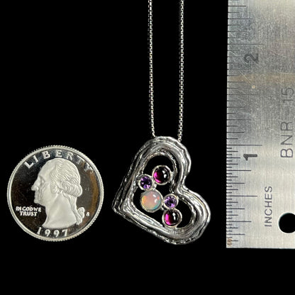 A silver heart necklace set with opal, amethysts, and rhodolite garnets, shown next to a ruler and quarter.