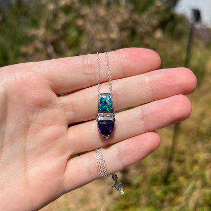 Hand holding a sterling silver opal inlay, white CZ, and purple trillion cut CZ drop necklace in natural light against blurred outdoor backdrop.