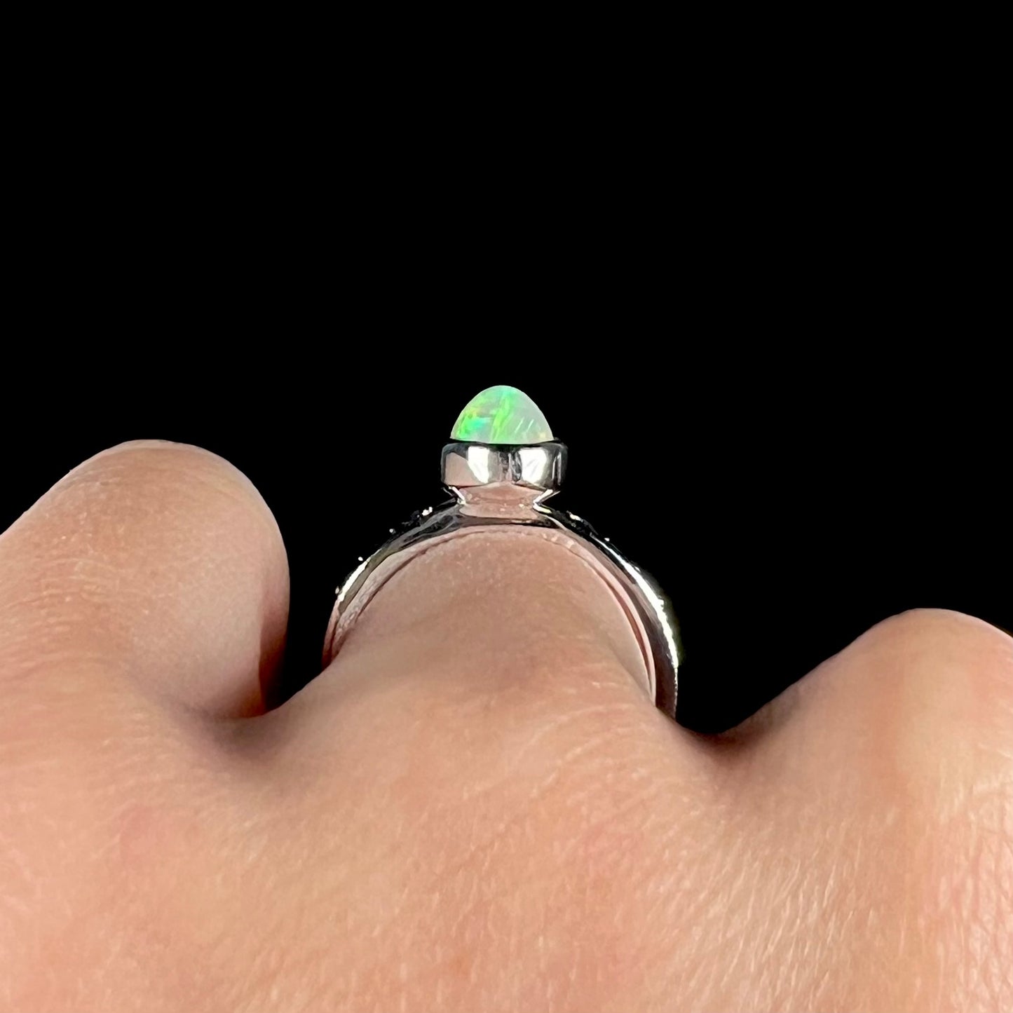 Macro side gallery view of a silver opal ring worn on a hand against a black background.