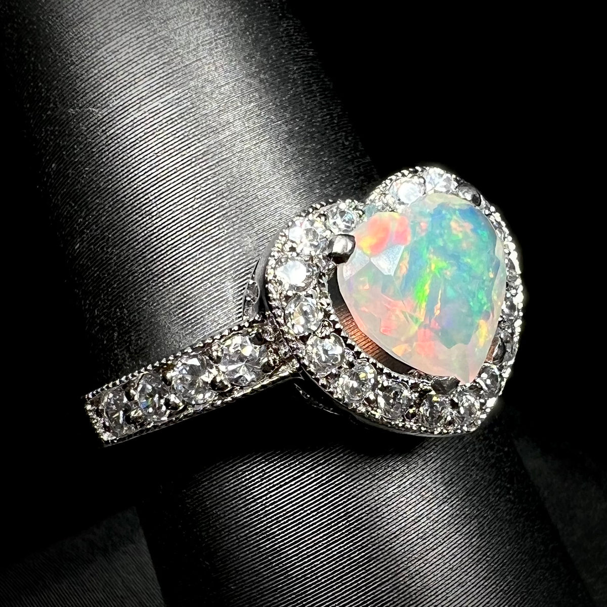 Women's sterling silver heart shaped opal ring with CZ halo design and milgrain details, shown close-up from angle on ring display