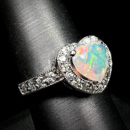Women's sterling silver heart shaped opal ring with CZ halo design and milgrain details, shown close-up from angle on ring display