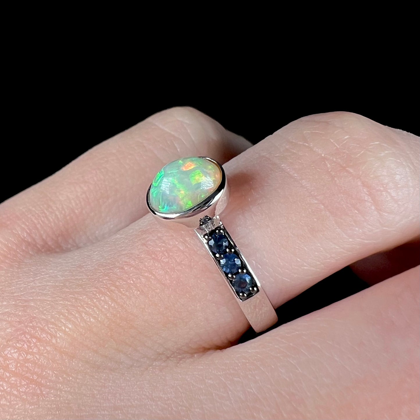 Macro view of a silver opal ring with dark blue topaz accents worn on a finger, shown at an angle.