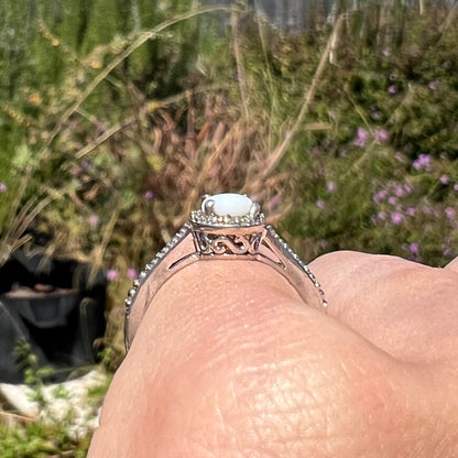 Side profile view of a silver opal ring with filigree design along its basket, worn on a finger in natural light.
