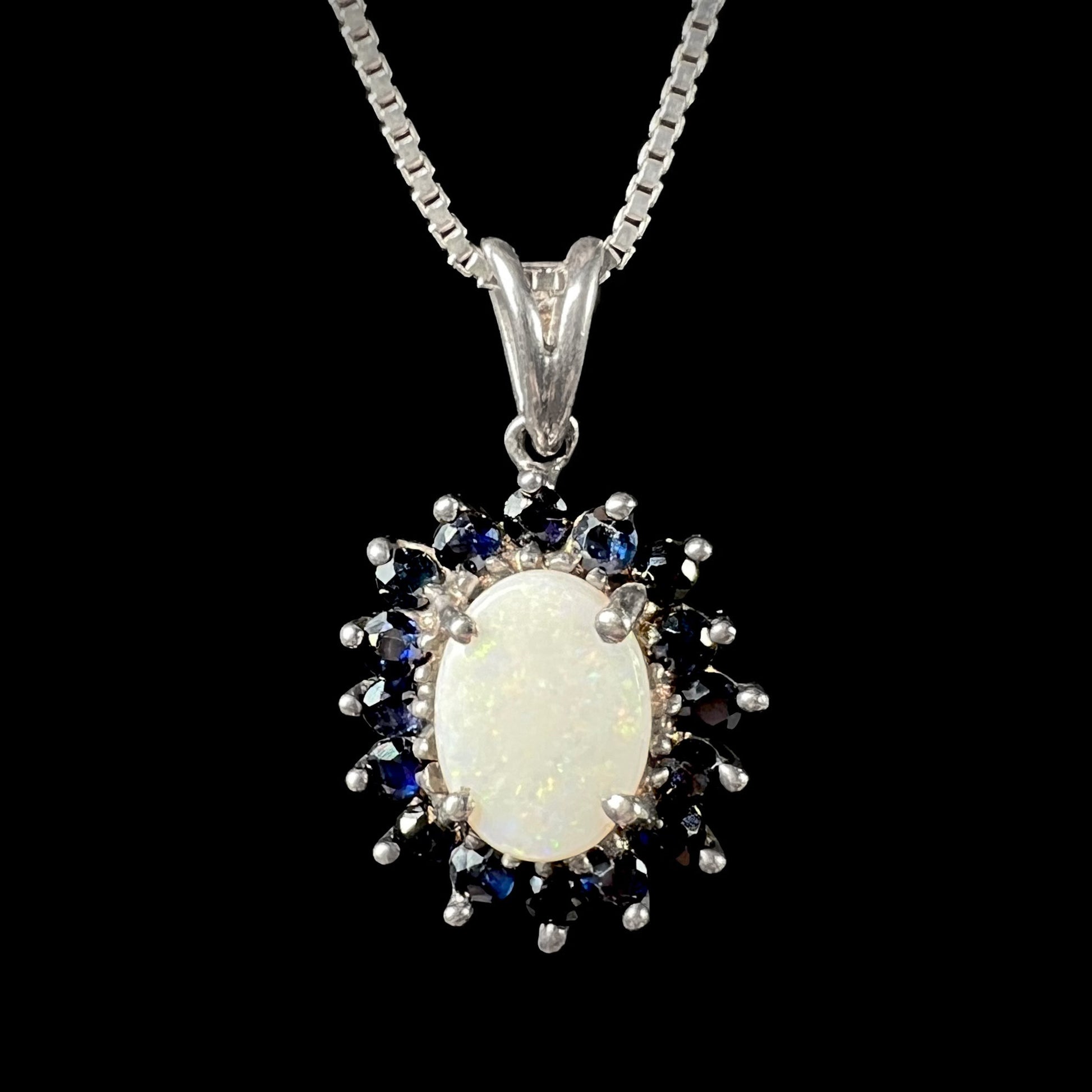 A sterling silver opal and sapphire halo necklace displayed against a black background, showing subtle yellow pinfire flashes.