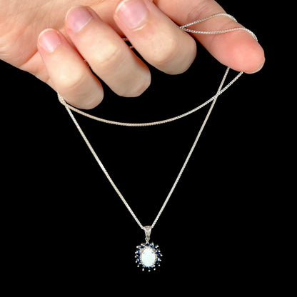 A sterling silver opal and sapphire halo necklace being dangled by its chain in a hand against a black background.