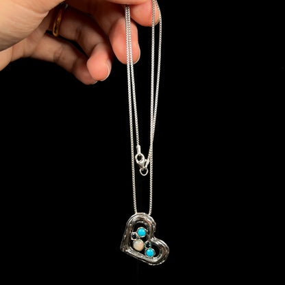 A sterling silver heart necklace set with Ethiopian opal, turquoise, and blue topaz, dangled from its chain by a hand.