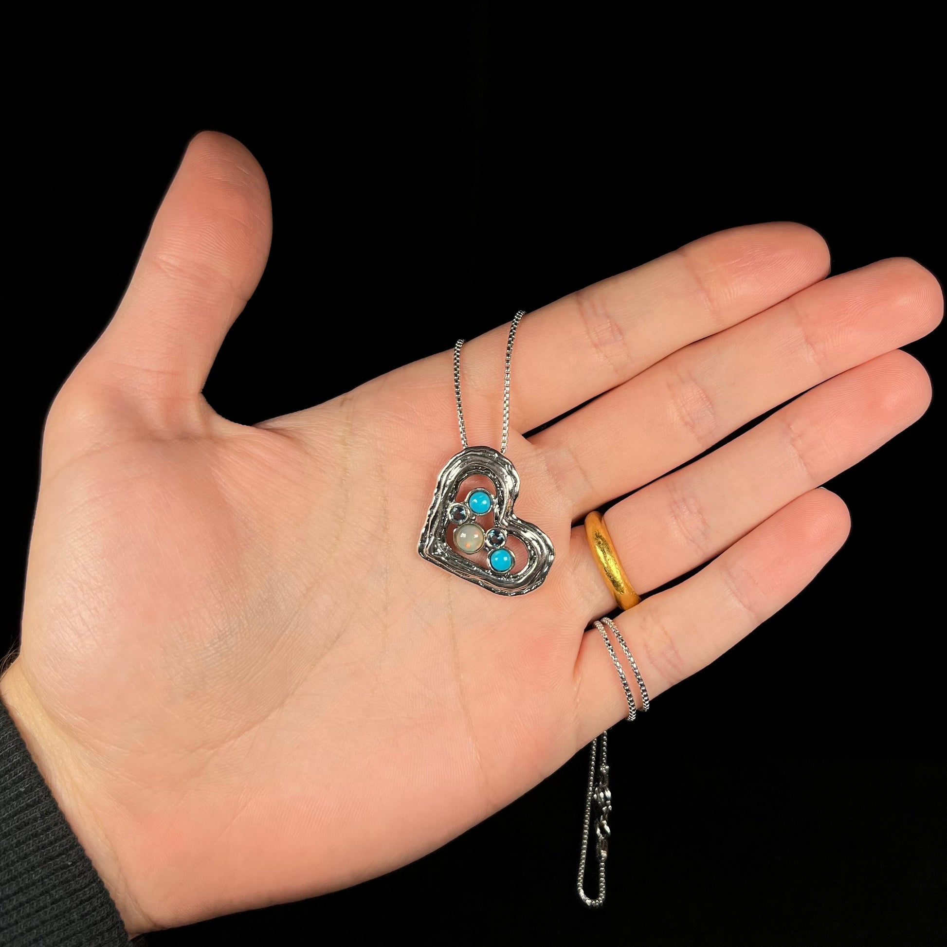 A sterling silver heart necklace set with opal, turquoise, and blue topaz, shown in a hand against black background.