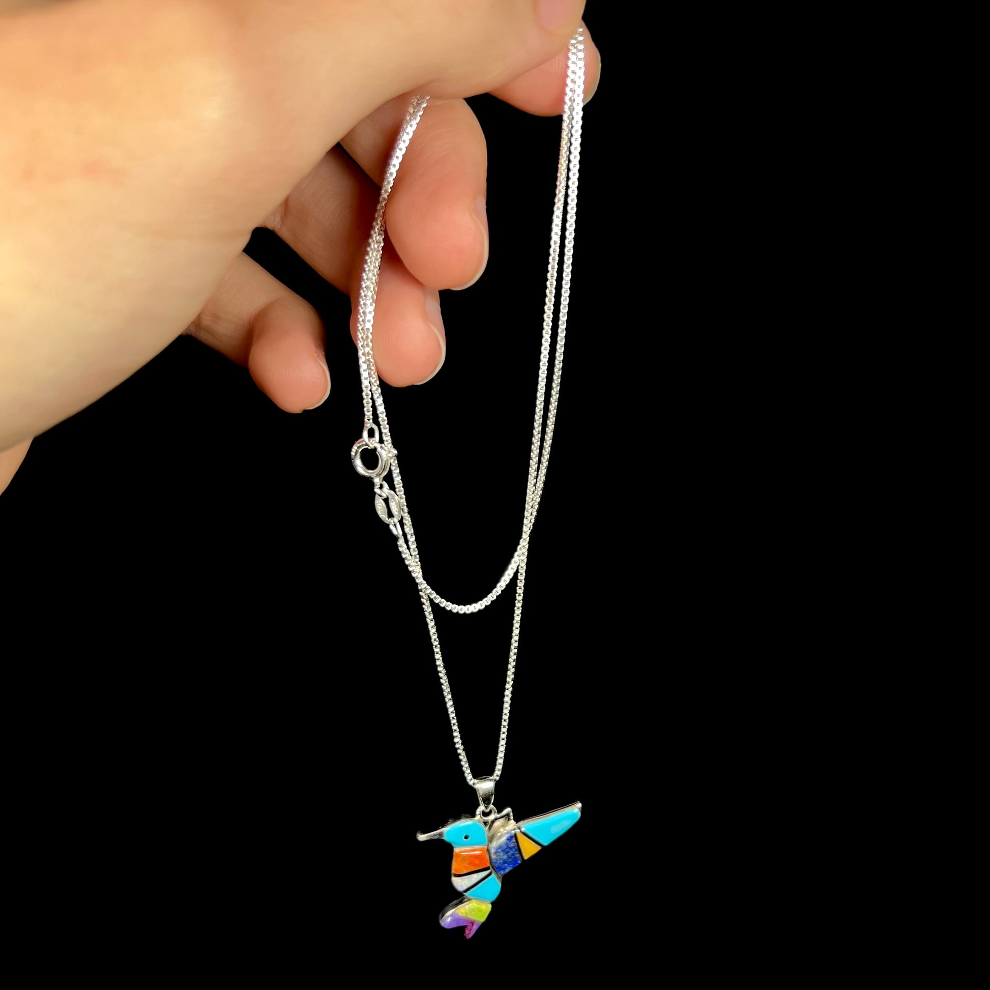 A multi-stone inlay hummingbird necklace in sterling silver dangled from a hand by the chain.