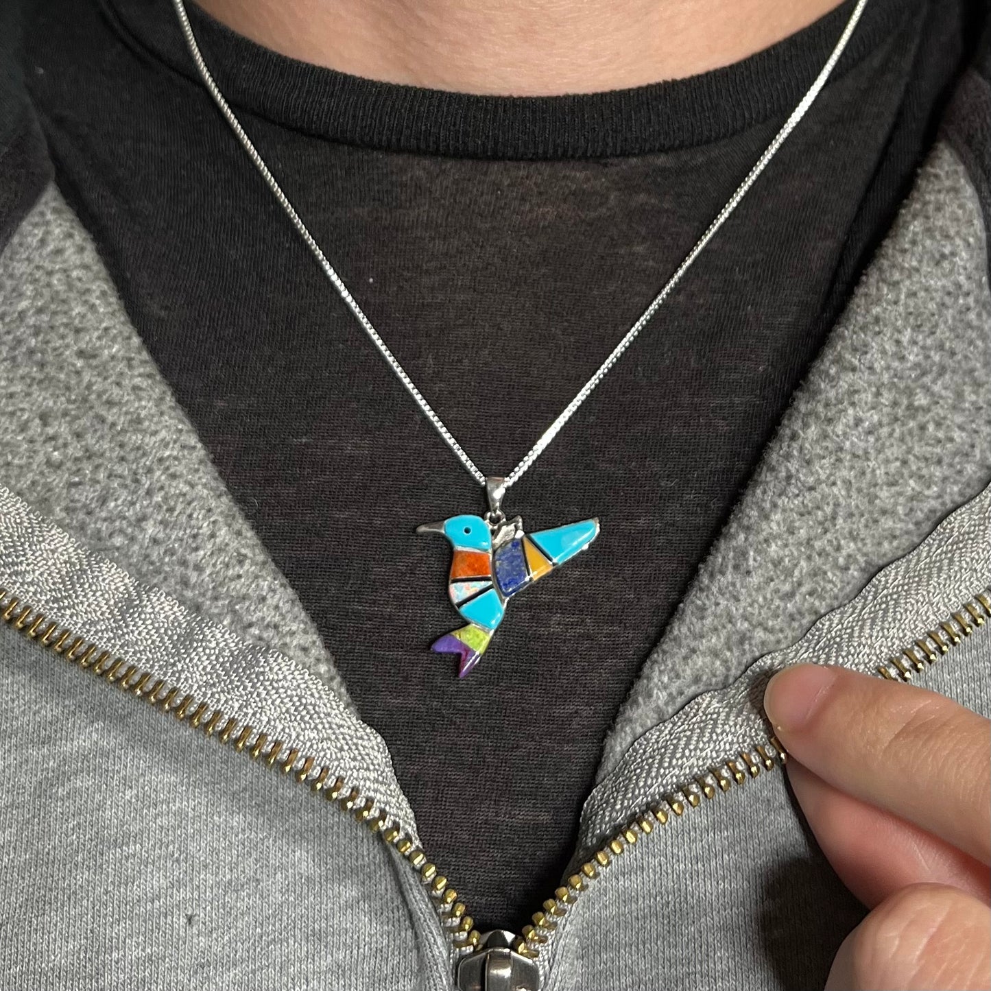 A multi-stone inlay hummingbird necklace in sterling silver, worn arond a neck against a dark shirt.