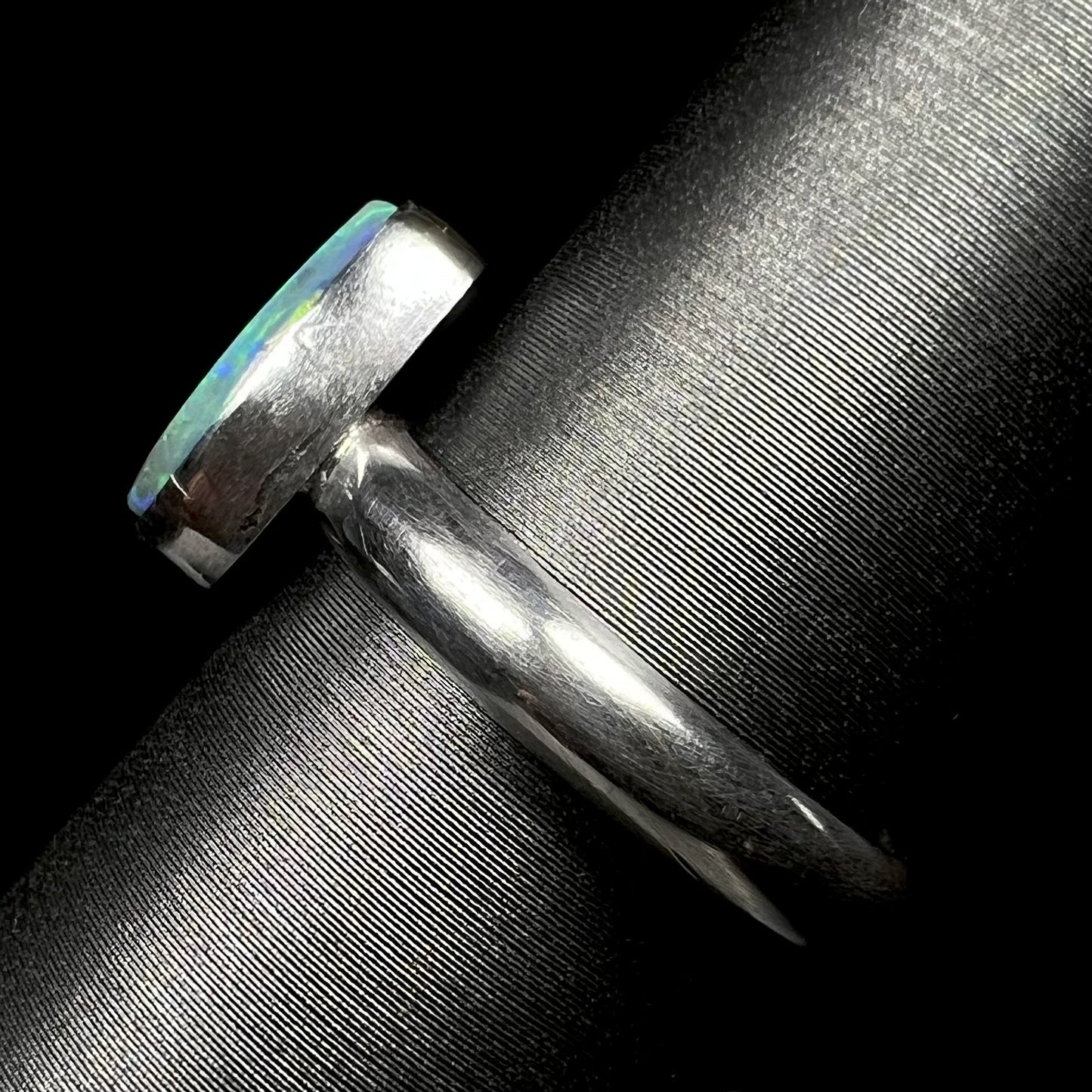 Side view of a simple sterling silver solitaire ring set with an opal, shown on a ring display.  The ring's shank and bezel setting are in focus.