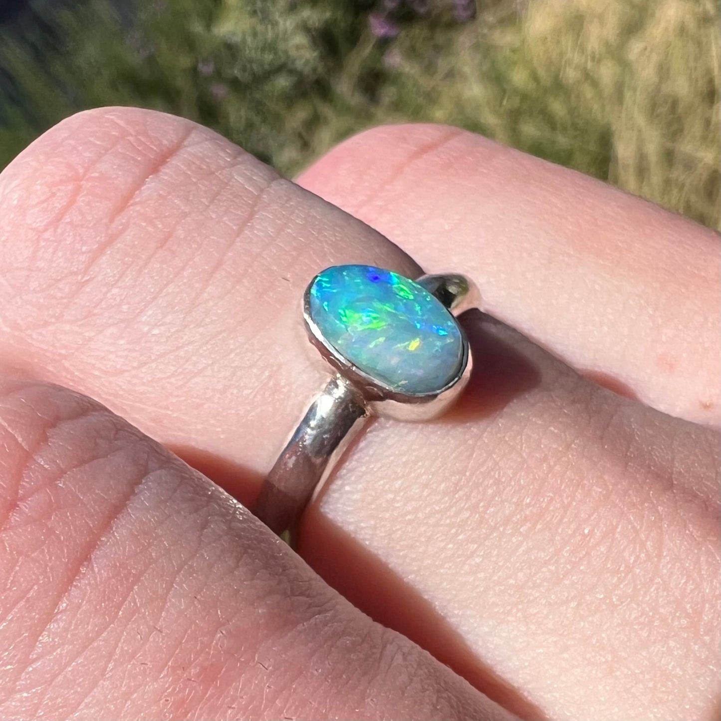 A simple women's blue and green opal ring made from sterling silver shown close-up from an angle while worn on a finger in sunlight.