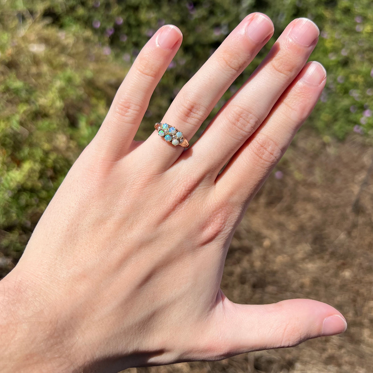 Sunlit hand wearing a Victorian six-stone opal ring with pearl accents in gold on ring finger against blurred outdoor background