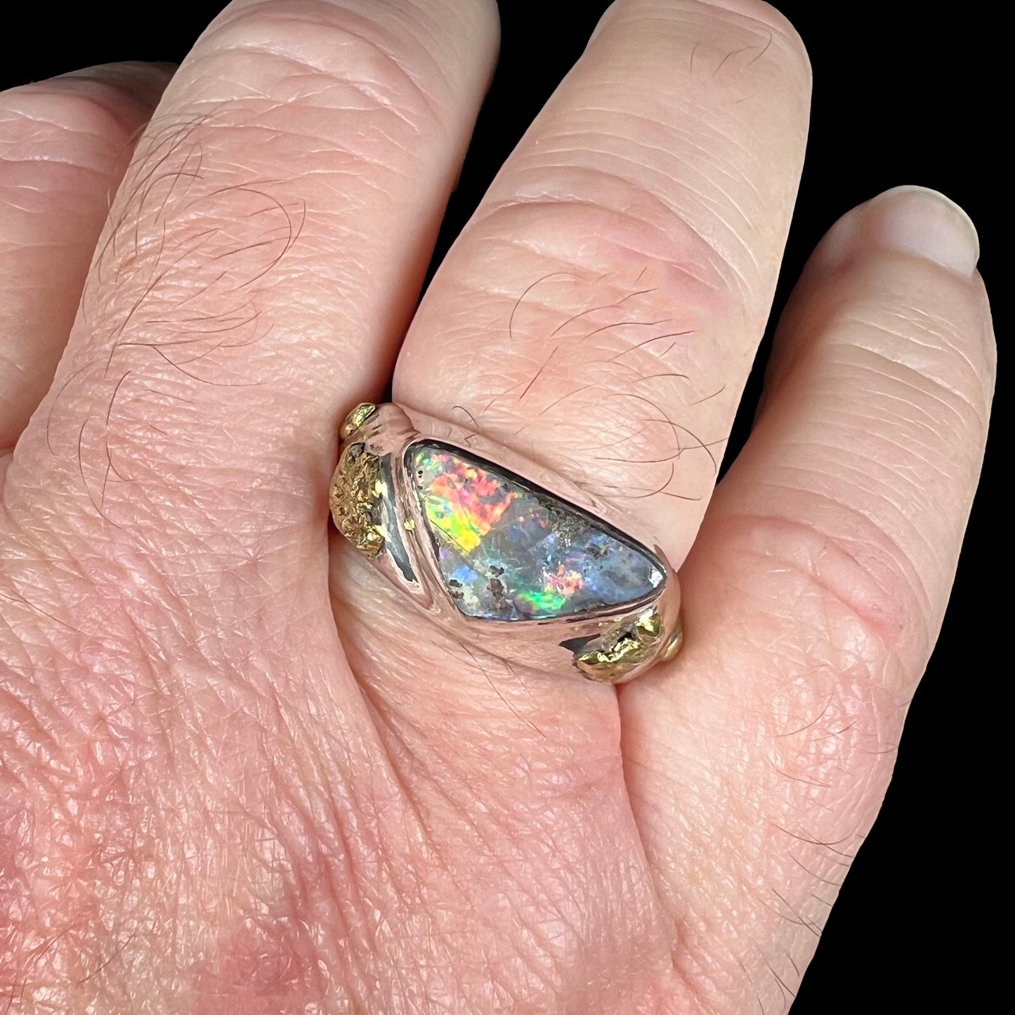 Close-up view of a size 12 sterling silver boulder opal ring worn on a man's finger against a black backdrop.  The opal has red, orange, and flashes in studio light.
