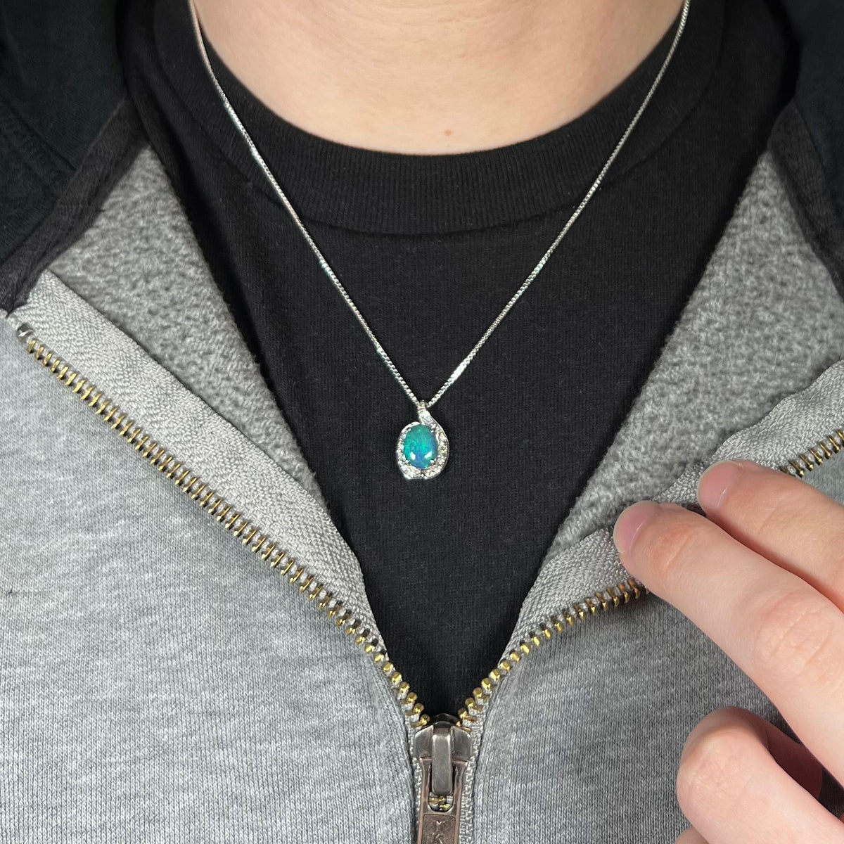 Close-up of person's chest and neck wearing small women's pendant necklace set with Australian black opal in asymmetrical halo design, worn against dark shirt.