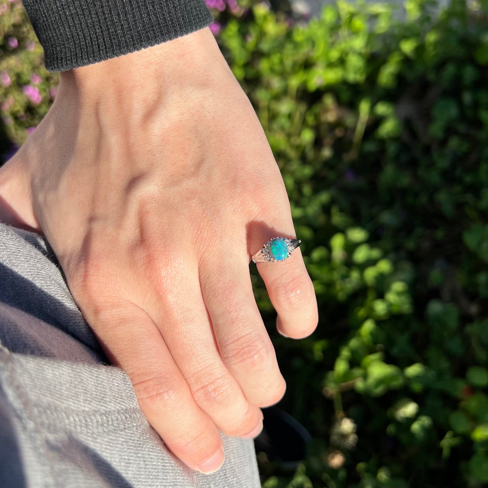 A small, delicate black opal platinum ring worn on a hand in the sunlight.