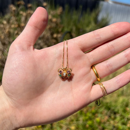 Hand holding a small gold opal and gemstone butterfly necklace in natural sunlight against blurred green backdrop.
