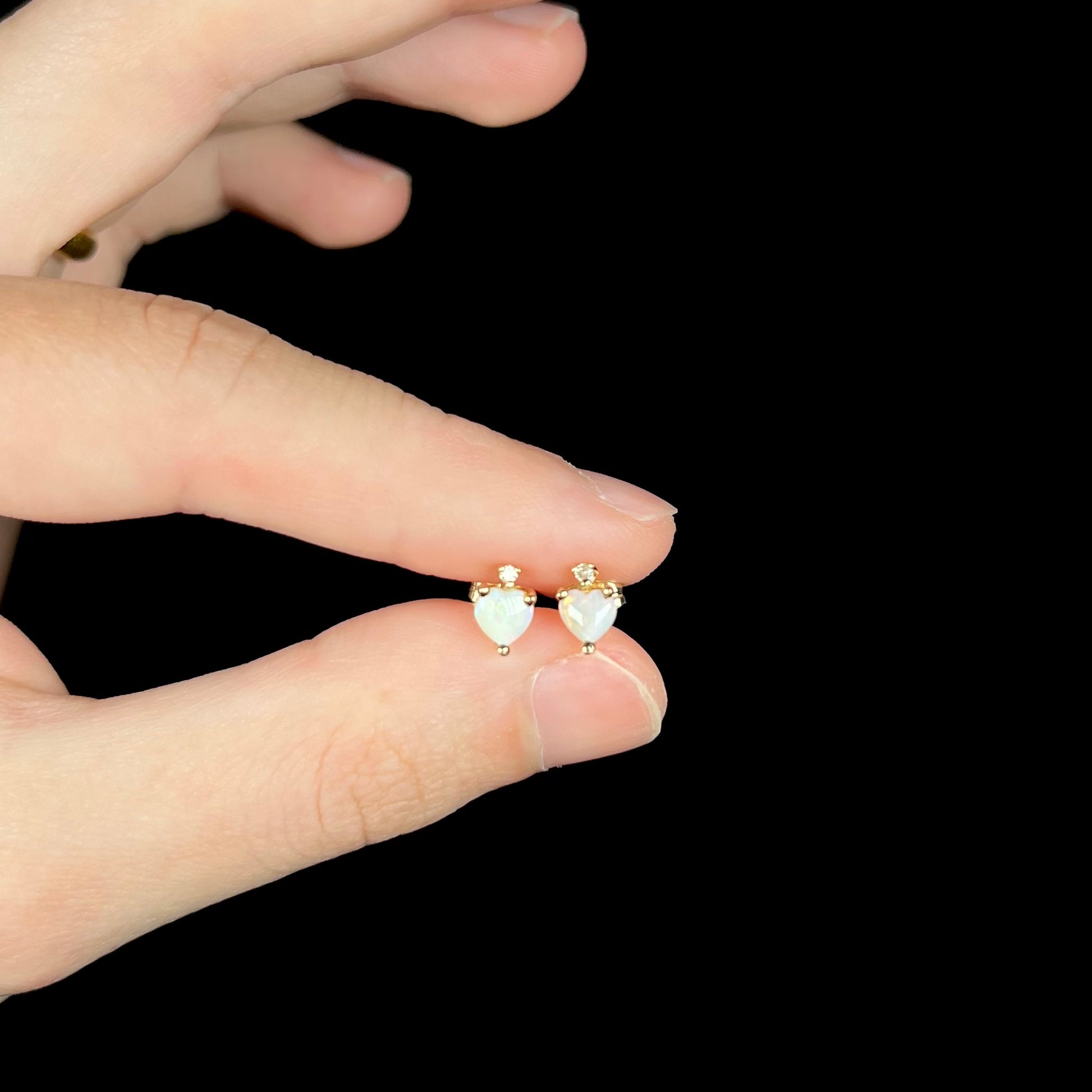 Pair of small heart shaped opal and diamond stud earrings in 14k yellow gold, held between fingers.