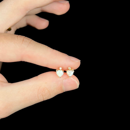 Pair of small heart shaped opal and diamond stud earrings in 14k yellow gold, held between fingers.