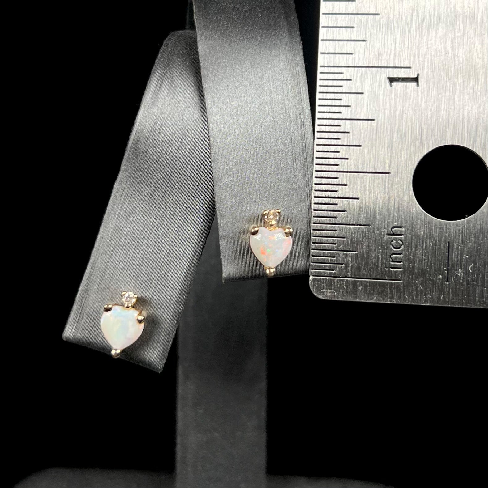 A pair of small opal heart stud earrings measuring 0.3 inches long, shown on earring display next to ruler for scale.