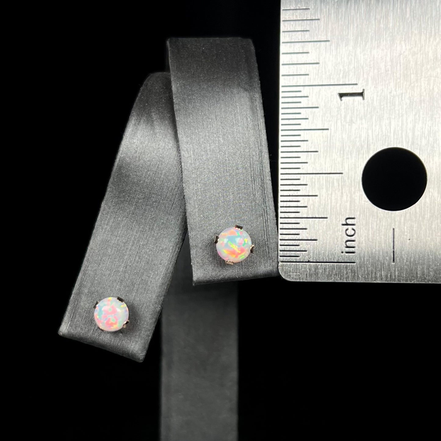 A pair of small lab-created opal stud earrings in 14k gold, shown on an earring display next to metal ruler.  The earrings measure under one-quarter of an inch in diameter.