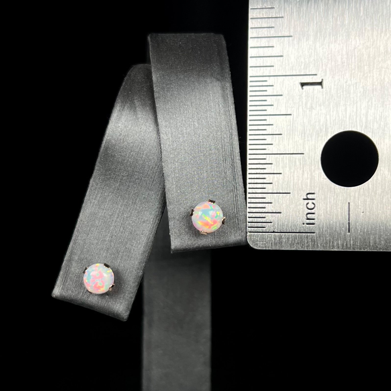 A pair of small lab-created opal stud earrings in 14k gold, shown on an earring display next to metal ruler.  The earrings measure under one-quarter of an inch in diameter.