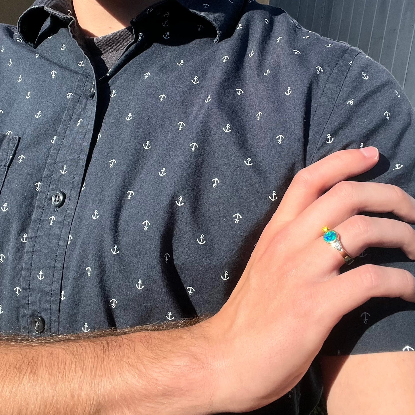 A hand modeling a small, round boulder opal and diamond ring as it rests on the model's opposing upper arm.  The diamonds sparkle in sunlight.