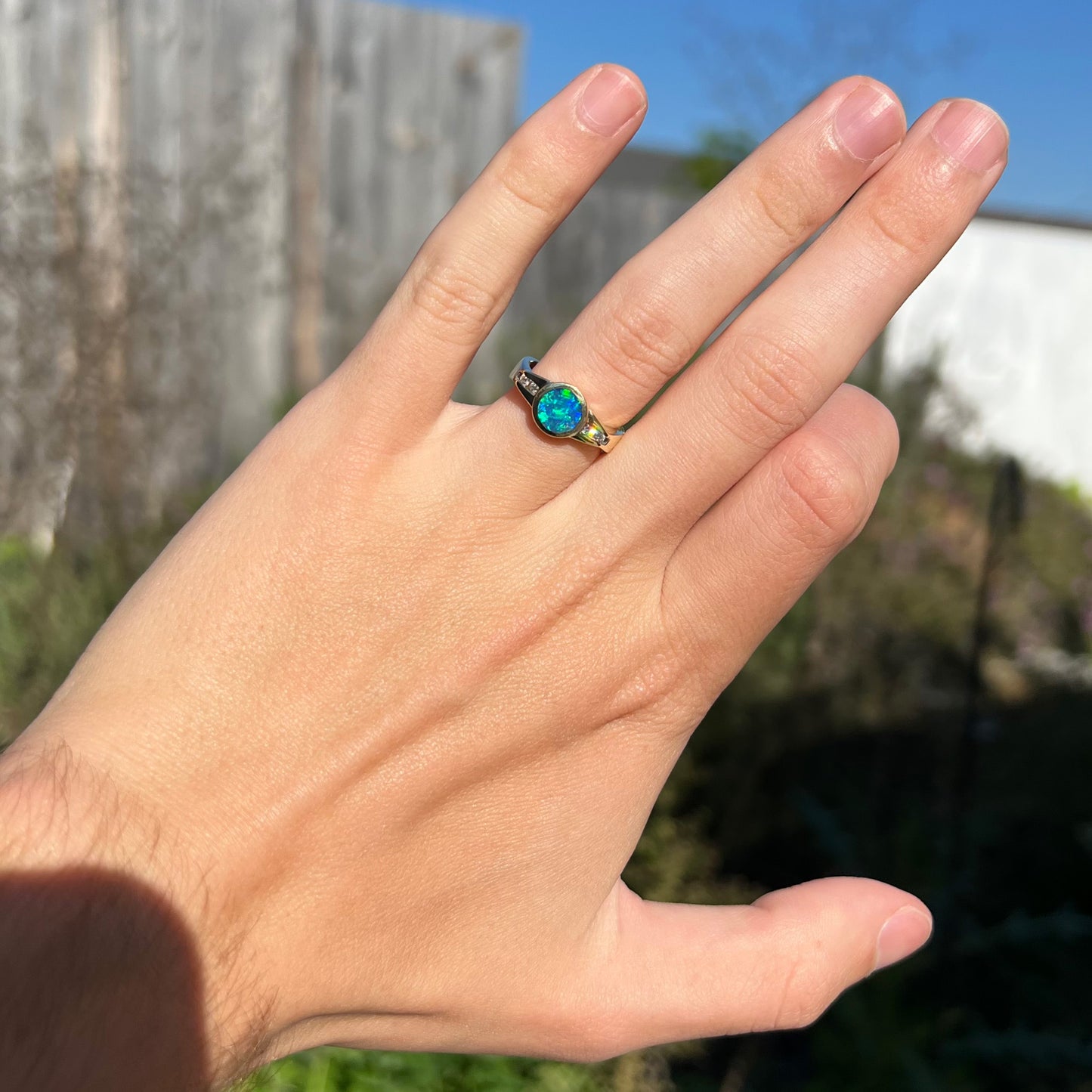 A small, round boulder opal ring with diamond accents, worn on a hand, shown against an outdoor background.  The diamonds sparkle in the sunlight.