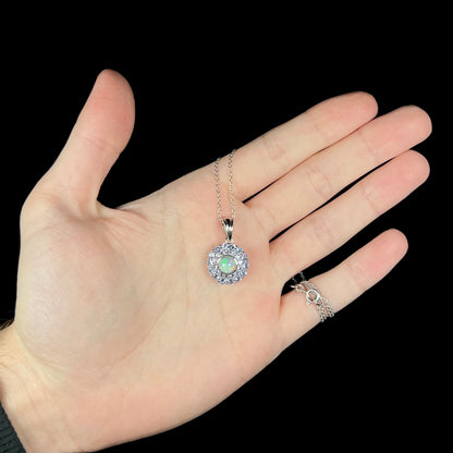 Hand holding a small, round women's Ethiopian opal and tanzanite necklace under LED studio lights, its silver chain wrapping around the hand's pinky finger.