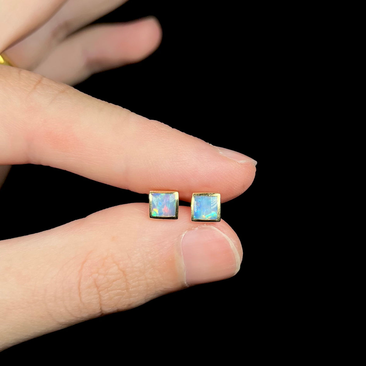 Fingers holding small square cut opal stud earrings in 14k gold