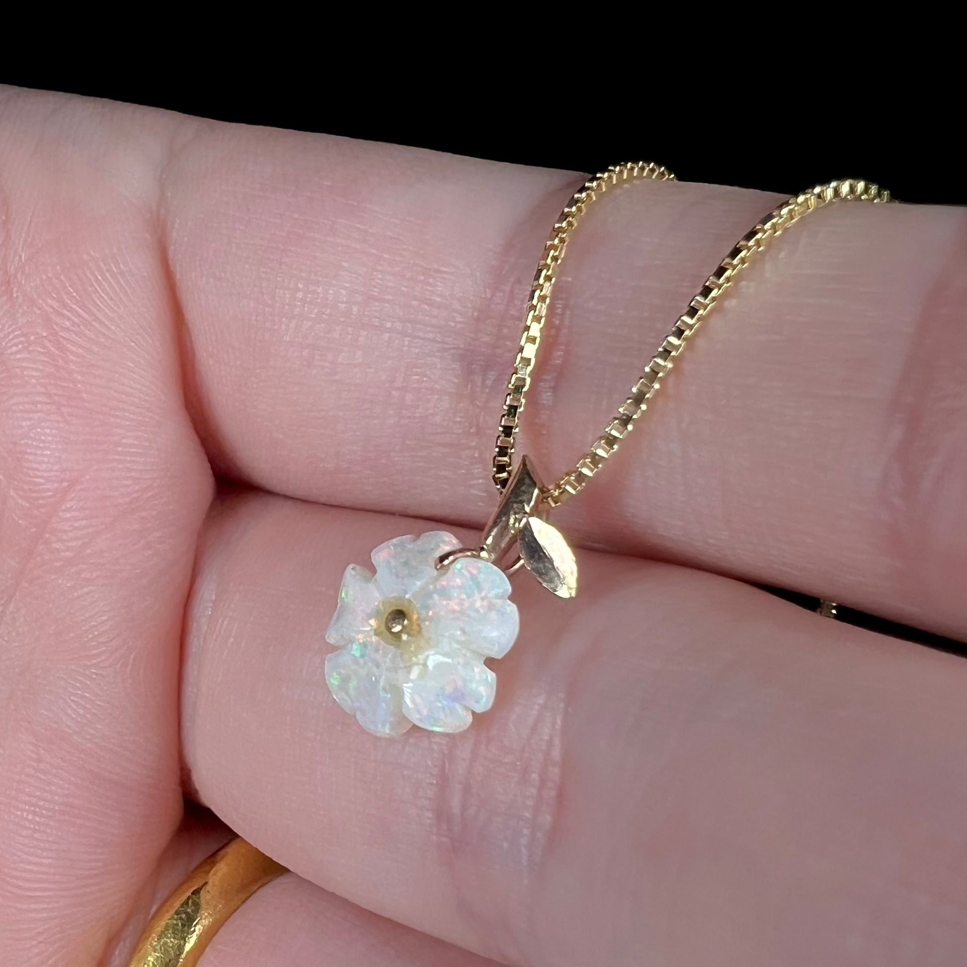 A small white opal flower pendant shown close-up in a hand from an angle on a box chain under LED light against a black backdrop.