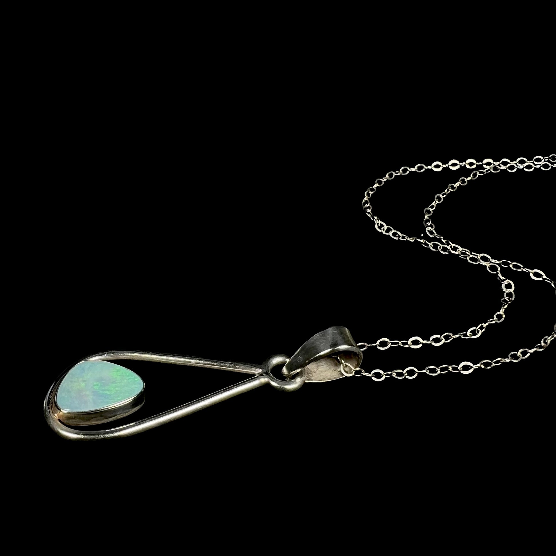 A sterling silver opal doublet necklace shown laying on a black background.  The opal has soft green play of color under cool studio light.
